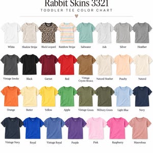 May include: A Rabbit Skins 3321 toddler tee color chart displaying thirty-two short-sleeve t-shirts in various solid colors, stripes, and patterns. Each shirt is labeled with its specific color name, including white, black, red, and navy.