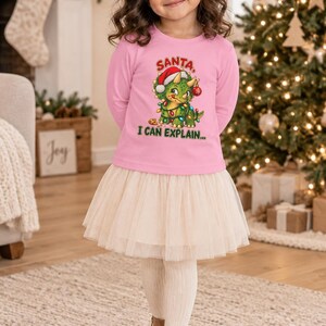 K&ouml;nnte beinhalten: Ein lang&auml;rmeliges rosa Shirt mit einer Grafik eines gr&uuml;nen Triceratops, der eine Weihnachtsm&uuml;tze tr&auml;gt und in Lichterketten gewickelt ist, mit dem Text "SANTA I CAN EXPLAIN..." und einem cremefarbenen T&uuml;llrock kombiniert.