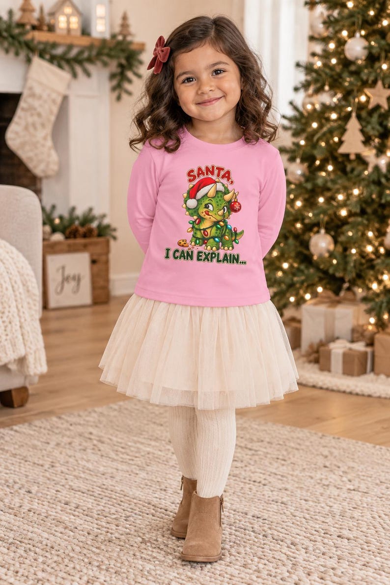 K&ouml;nnte beinhalten: Ein lang&auml;rmeliges rosa Shirt mit einer Grafik eines gr&uuml;nen Triceratops, der eine Weihnachtsm&uuml;tze tr&auml;gt und in Lichterketten gewickelt ist, mit dem Text "SANTA I CAN EXPLAIN..." und einem cremefarbenen T&uuml;llrock kombiniert.
