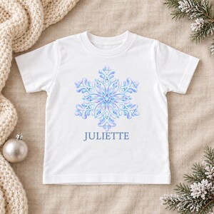 May include: A white short-sleeved t-shirt featuring a large, intricate, light blue and purple snowflake graphic centered on the chest. Below the snowflake, the name JULIETTE is printed in a simple, clear, light blue sans-serif font.