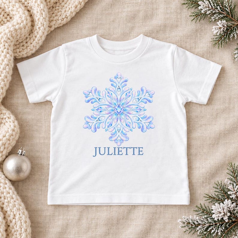May include: A white short-sleeved t-shirt featuring a large, intricate, light blue and purple snowflake graphic centered on the chest. Below the snowflake, the name JULIETTE is printed in a simple, clear, light blue sans-serif font.