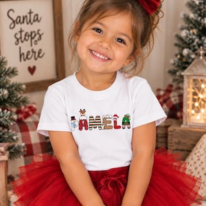 May include: A white short-sleeved t-shirt featuring the name PAMELA printed in colorful, festive holiday-themed letters, including a snowman, reindeer, Santa Claus, and elf patterns, paired with a layered red tulle skirt with a bow.