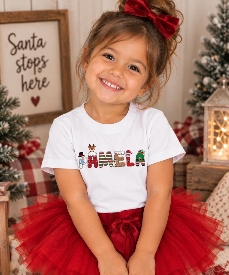 May include: A white short-sleeved t-shirt featuring the name PAMELA printed in colorful, festive holiday-themed letters, including a snowman, reindeer, Santa Claus, and elf patterns, paired with a layered red tulle skirt with a bow.