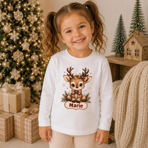 May include: A white long-sleeve shirt featuring a graphic of a cartoon reindeer with a plaid bow and the name Marie printed below. The reindeer is surrounded by small snowflakes and pine branches, centered on the front of the shirt.