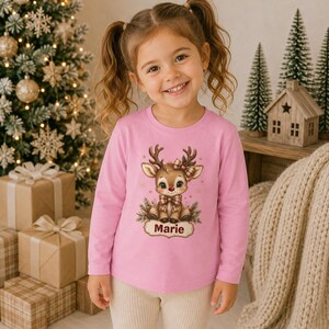 May include: A light pink long-sleeve shirt featuring a graphic of a cartoon reindeer with a plaid bow and scarf, surrounded by pine branches and gold stars. Below the reindeer is a decorative label with the name Marie printed in dark red text.