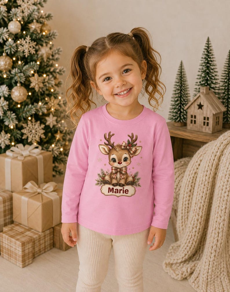 May include: A light pink long-sleeve shirt featuring a graphic of a cartoon reindeer with a plaid bow and scarf, surrounded by pine branches and gold stars. Below the reindeer is a decorative label with the name Marie printed in dark red text.