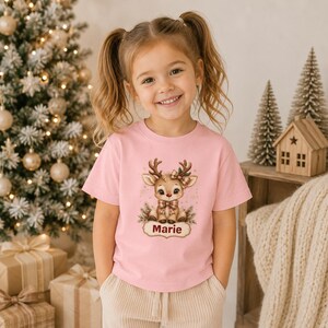 May include: A light pink short-sleeved t-shirt featuring a graphic of a cartoon reindeer with a plaid bow and scarf. Below the reindeer, a decorative label contains the name Marie in dark red text, surrounded by small star accents.