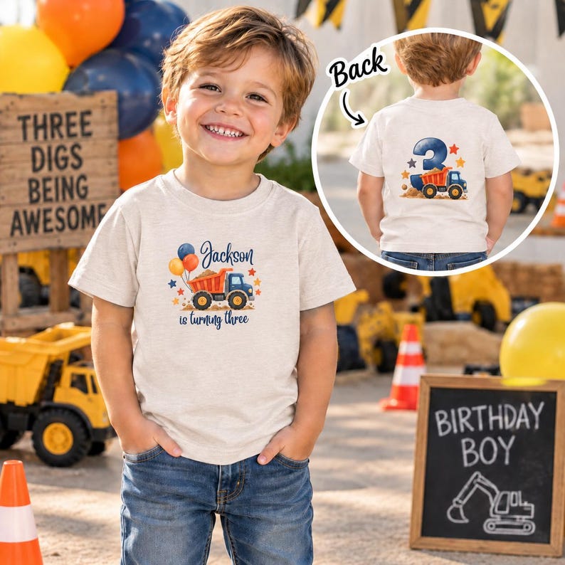 May include: A beige short-sleeve t-shirt featuring a graphic of a dump truck with balloons and the text Jackson is turning three on the front, and a large number three with a dump truck graphic on the back, shown in a circular inset.