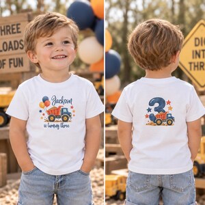 May include: A white short-sleeved t-shirt featuring a graphic of a dump truck, balloons, and the text Jackson is turning three on the front, and a large number three with a dump truck graphic on the back, paired with blue denim jeans.