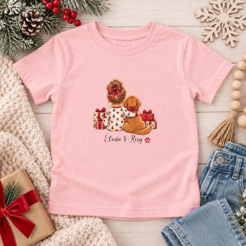 May include: A light pink short-sleeved t-shirt featuring a graphic of a child with a red bow and a golden dog sitting together surrounded by Christmas presents. The text "Elonie & Rory" is printed below the illustration in a cursive font.