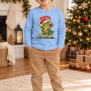 K&ouml;nnte beinhalten: Ein hellblaues Langarmshirt mit einer Grafik eines gr&uuml;nen Triceratops, der eine Weihnachtsm&uuml;tze tr&auml;gt und in bunte Lichterketten gewickelt ist. Der Text &uuml;ber dem Dinosaurier lautet SANTA und darunter I CAN EXPLAIN... in gr&uuml;nen Buchstaben.