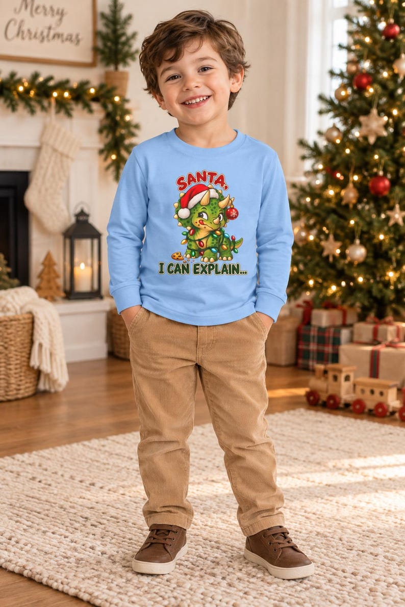 K&ouml;nnte beinhalten: Ein hellblaues Langarmshirt mit einer Grafik eines gr&uuml;nen Triceratops, der eine Weihnachtsm&uuml;tze tr&auml;gt und in bunte Lichterketten gewickelt ist. Der Text &uuml;ber dem Dinosaurier lautet SANTA und darunter I CAN EXPLAIN... in gr&uuml;nen Buchstaben.