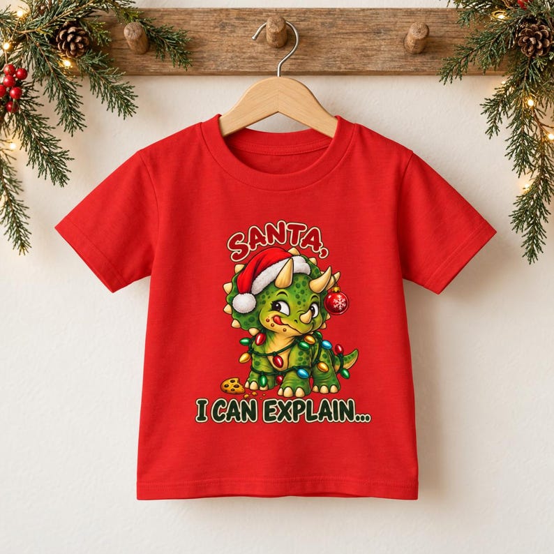 K&ouml;nnte beinhalten: Ein leuchtend rotes Kurzarm-T-Shirt mit einer Grafik eines gr&uuml;nen Triceratops, der eine Weihnachtsmannm&uuml;tze tr&auml;gt und in bunte Lichterketten gewickelt ist. Der Dinosaurier h&auml;lt eine rote Kugel und der Text lautet SANTA, I CAN EXPLAIN...