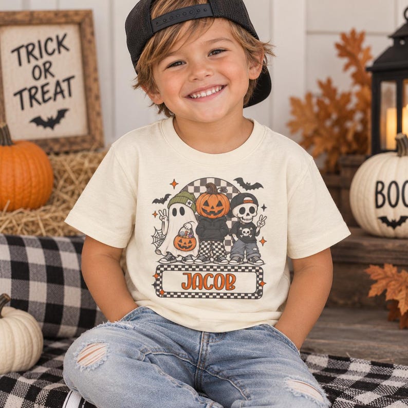 May include: A cream-colored short-sleeve t-shirt featuring a graphic print of a ghost, a pumpkin-headed figure, and a skeleton. The design includes bats, a spiderweb, and a checkered pattern. The name JACOB is printed in a rectangular box.