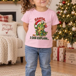 K&ouml;nnte beinhalten: Ein hellrosa kurz&auml;rmeliges T-Shirt mit einer Grafik eines gr&uuml;nen Triceratops, der eine Weihnachtsm&uuml;tze tr&auml;gt und in bunte Lichterketten gewickelt ist, mit dem Text SANTA, I CAN EXPLAIN in roten und gr&uuml;nen Buchstaben dar&uuml;ber und darunter.