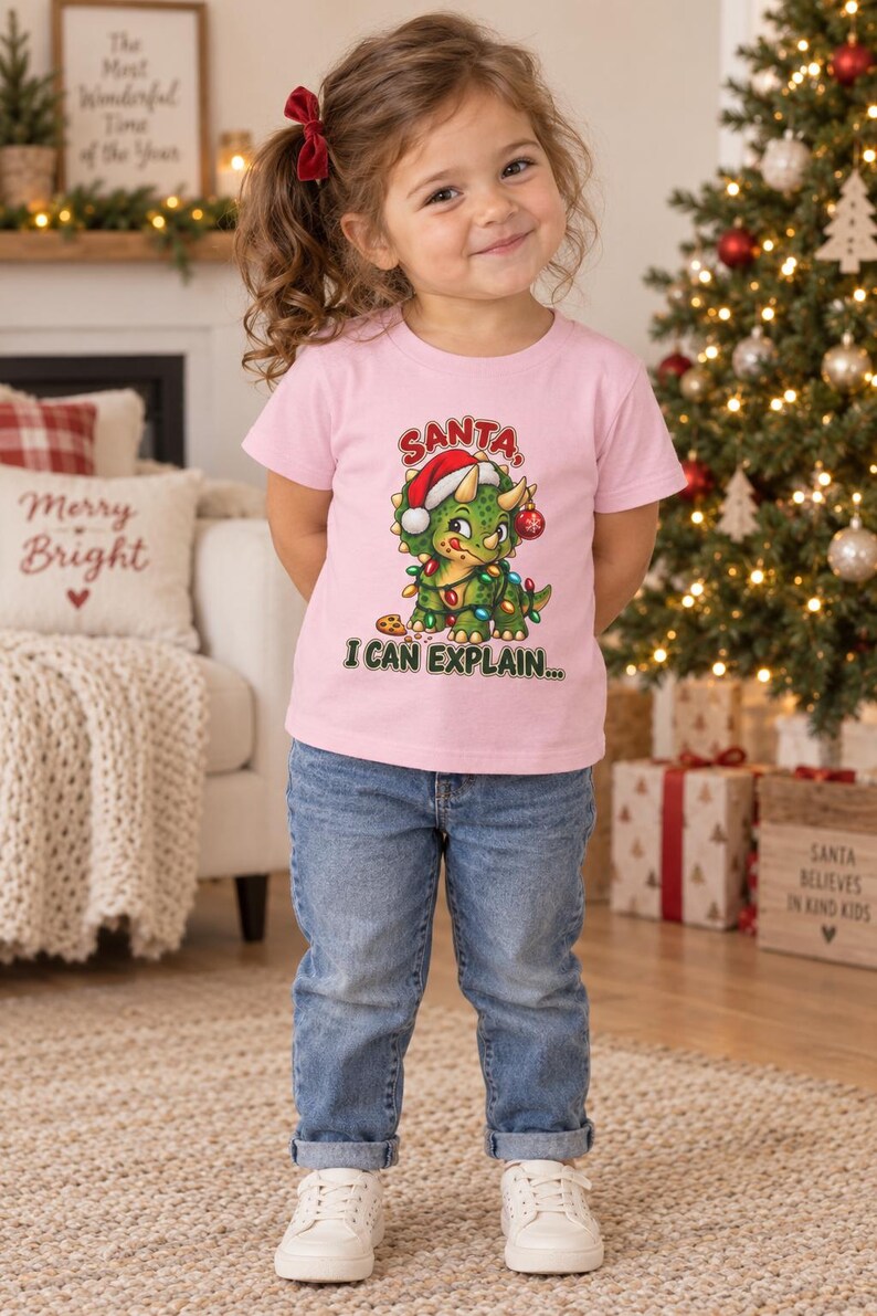 K&ouml;nnte beinhalten: Ein hellrosa kurz&auml;rmeliges T-Shirt mit einer Grafik eines gr&uuml;nen Triceratops, der eine Weihnachtsm&uuml;tze tr&auml;gt und in bunte Lichterketten gewickelt ist, mit dem Text SANTA, I CAN EXPLAIN in roten und gr&uuml;nen Buchstaben dar&uuml;ber und darunter.