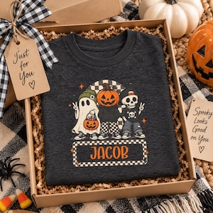 May include: A dark gray t-shirt featuring a graphic of a ghost, a pumpkin-headed figure, and a skeleton character. The design includes the name JACOB in orange text within a checkered border. The shirt is presented in a gift box with a tag reading Just for You.