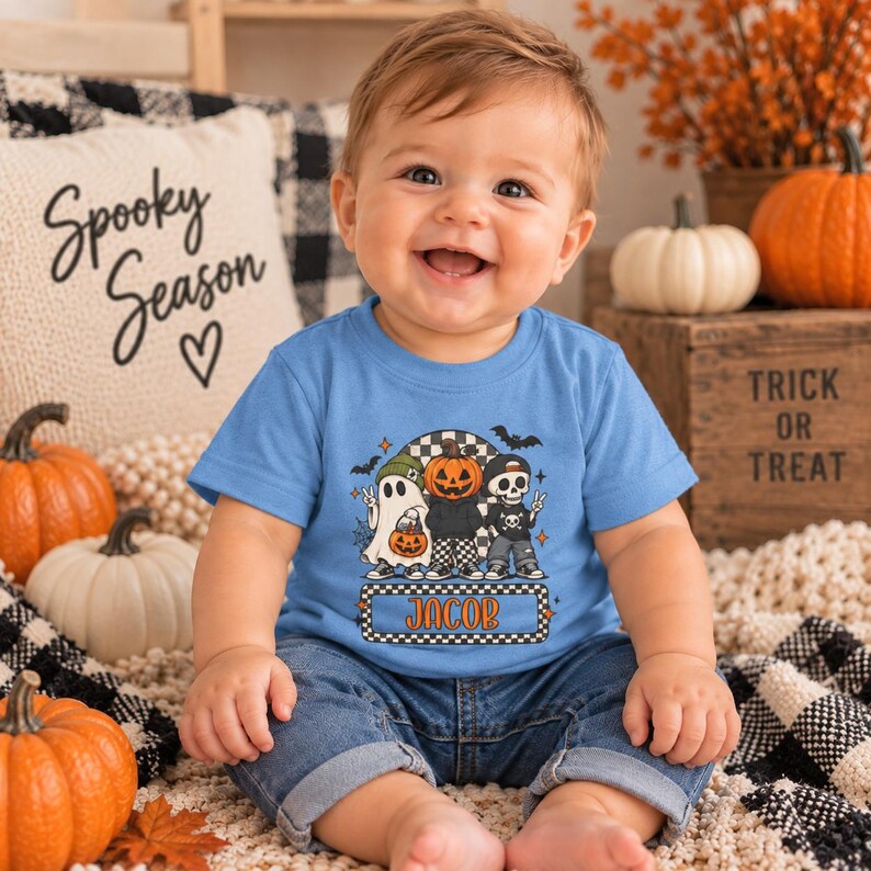May include: A light blue short-sleeve t-shirt featuring a graphic print of a ghost, a pumpkin-headed character, and a skeleton. The design includes the name JACOB in orange letters inside a checkered rectangular border with bats and stars.