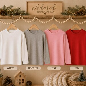 May include: Four long-sleeve crew neck shirts hanging on wooden hangers from a wooden rack. The shirts are solid white, heather gray, light pink, and bright red. Below each shirt is a label reading White, Heather, Pink, and Red respectively.