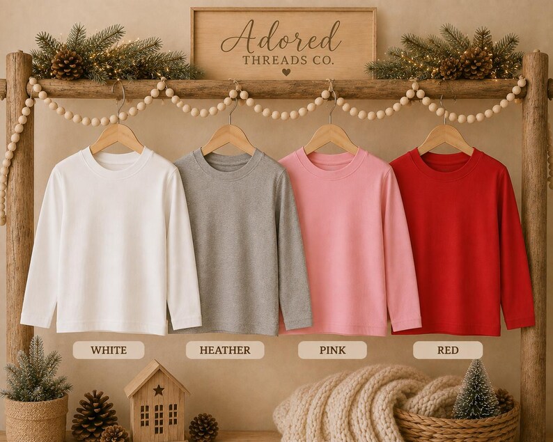 May include: Four long-sleeve crew neck shirts hanging on wooden hangers from a wooden rack. The shirts are solid white, heather gray, light pink, and bright red. Below each shirt is a label reading White, Heather, Pink, and Red respectively.