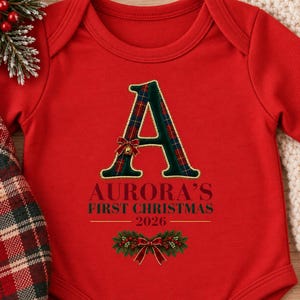 May include: A bright red long-sleeve baby bodysuit featuring a large letter A with a green and red plaid pattern. Below the letter, the text reads AURORA'S FIRST CHRISTMAS 2026 in dark green and red, accented by a festive pine and red bow graphic.