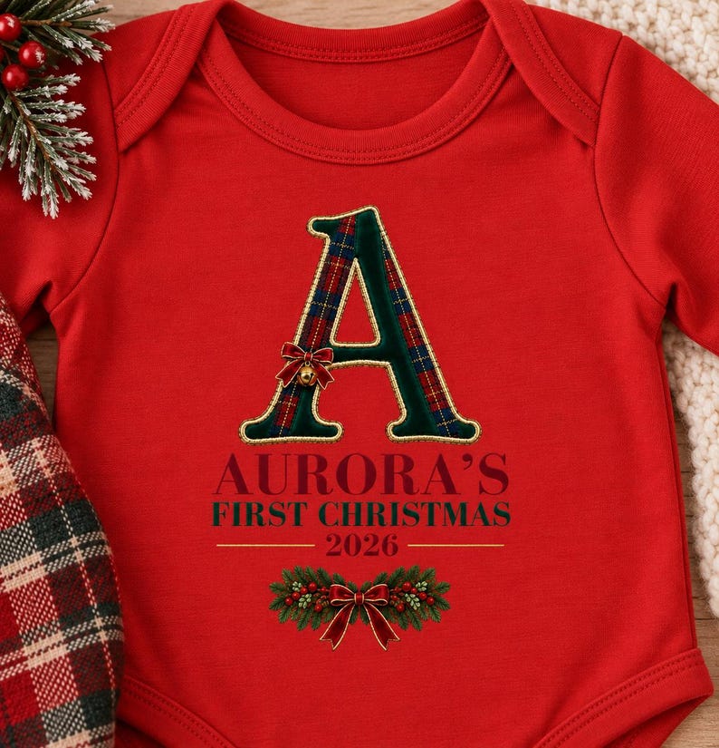May include: A bright red long-sleeve baby bodysuit featuring a large letter A with a green and red plaid pattern. Below the letter, the text reads AURORA'S FIRST CHRISTMAS 2026 in dark green and red, accented by a festive pine and red bow graphic.