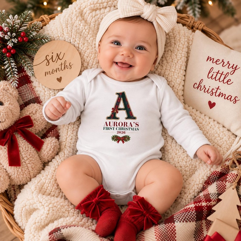 May include: A white long-sleeve baby bodysuit featuring a plaid letter A, the text Aurora's First Christmas 2026, and a holly graphic. The baby wears a cream-colored bow headband and red socks with bows, surrounded by a wooden six months sign.