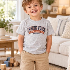 May include: A light gray short-sleeved t-shirt featuring the text INSIDE VOICE CURRENTLY UNAVAILABLE ETHAN AGE 3 in navy blue and orange lettering, paired with tan khaki shorts. The shirt has a crew neckline and a standard fit design.