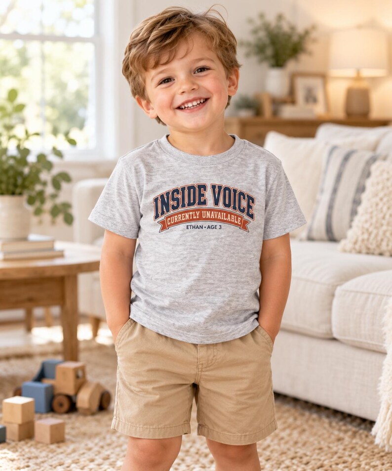 May include: A light gray short-sleeved t-shirt featuring the text INSIDE VOICE CURRENTLY UNAVAILABLE ETHAN AGE 3 in navy blue and orange lettering, paired with tan khaki shorts. The shirt has a crew neckline and a standard fit design.