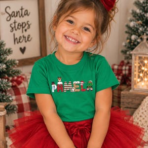 May include: A green short-sleeved t-shirt featuring the name PAMELA printed in a festive holiday font. Each letter is decorated with Christmas-themed graphics, including a snowman, a reindeer, a penguin, and an elf hat, paired with a red tulle skirt.