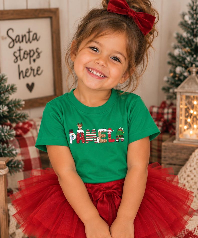 May include: A green short-sleeved t-shirt featuring the name PAMELA printed in a festive holiday font. Each letter is decorated with Christmas-themed graphics, including a snowman, a reindeer, a penguin, and an elf hat, paired with a red tulle skirt.