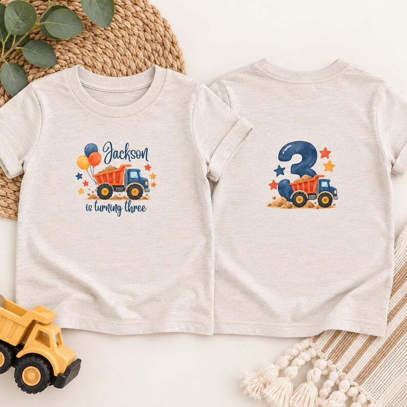May include: Two light heather gray t-shirts. The left shirt features a blue and orange dump truck, balloons, stars, and the text "Jackson is turning three". The right shirt displays a large blue number "3" with a dump truck and stars.