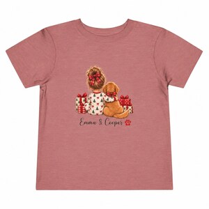 May include: A dusty rose short-sleeve t-shirt featuring a graphic of a child with a hair bow and a golden dog sitting together among Christmas presents. The text "Emma & Cooper" is printed below the illustration with a small red paw print icon.