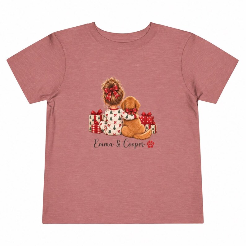 May include: A dusty rose short-sleeve t-shirt featuring a graphic of a child with a hair bow and a golden dog sitting together among Christmas presents. The text "Emma & Cooper" is printed below the illustration with a small red paw print icon.