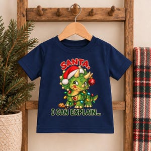 K&ouml;nnte beinhalten: Ein dunkelblaues Kurzarm-T-Shirt mit einer Grafik eines gr&uuml;nen Triceratops, der eine rote Weihnachtsmannm&uuml;tze tr&auml;gt, in bunte Lichterketten gewickelt ist und eine rote Kugel h&auml;lt. Der Text lautet SANTA, I CAN EXPLAIN... &uuml;ber und unter dem Dinosaurier.
