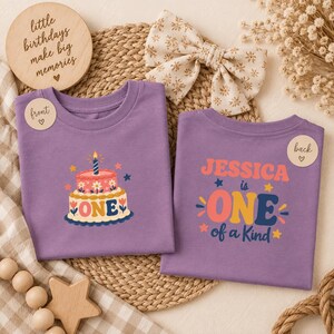 May include: Two purple t-shirts featuring birthday graphics. The front shirt displays a two-tier cake with a candle and the word ONE. The back shirt features the text JESSICA is ONE of a Kind with stars and decorative elements in pink, yellow, and blue.