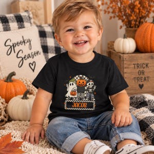 May include: A black short-sleeved t-shirt featuring a graphic print of a ghost, a pumpkin-headed character, and a skeleton. The design includes the name JACOB in orange text within a checkered rectangular border, set against a black background.