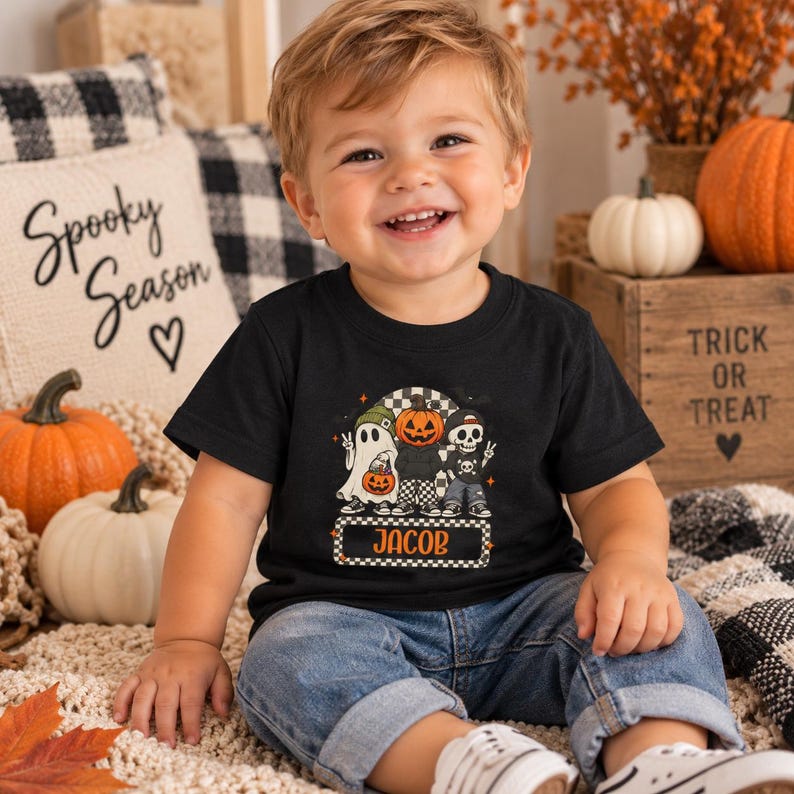 May include: A black short-sleeved t-shirt featuring a graphic print of a ghost, a pumpkin-headed character, and a skeleton. The design includes the name JACOB in orange text within a checkered rectangular border, set against a black background.
