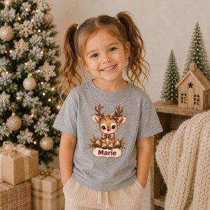 May include: A heather gray short-sleeved t-shirt featuring a graphic of a cartoon reindeer with a plaid bow and scarf. The reindeer is positioned above a decorative label containing the name Marie in dark red text, surrounded by pine branches.