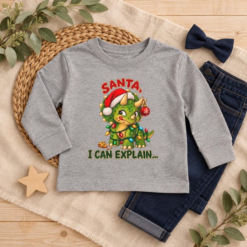 K&ouml;nnte beinhalten: Ein graues Langarmshirt mit einem gr&uuml;nen Triceratops, der eine Weihnachtsm&uuml;tze tr&auml;gt und in bunte Lichterketten gewickelt ist. Der Dinosaurier h&auml;lt eine rote Kugel und einen Keks. Der Text lautet SANTA, I CAN EXPLAIN in Rot und Gr&uuml;n.