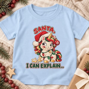 May include: A light blue t-shirt featuring a graphic of a cartoon unicorn wearing a red Santa hat and wrapped in colorful Christmas lights. The unicorn has a gold horn and holds a red ornament. The text reads SANTA, I CAN EXPLAIN...