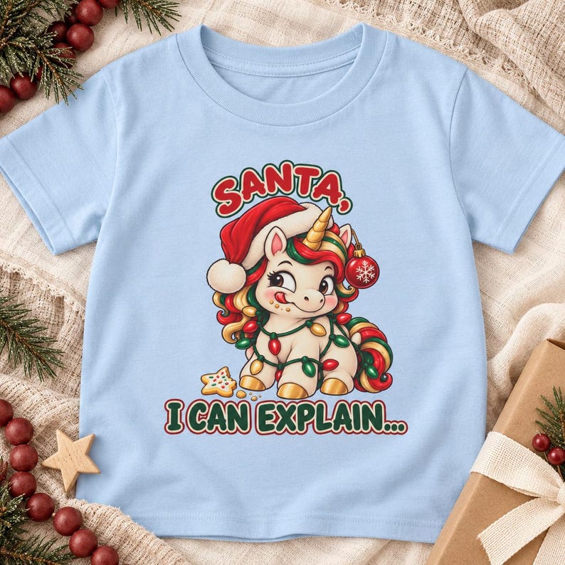 May include: A light blue t-shirt featuring a graphic of a cartoon unicorn wearing a red Santa hat and wrapped in colorful Christmas lights. The unicorn has a gold horn and holds a red ornament. The text reads SANTA, I CAN EXPLAIN...
