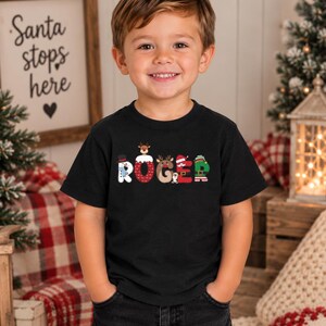 May include: A black short-sleeved t-shirt featuring the name ROGER printed in large, colorful letters. Each letter is decorated with Christmas-themed graphics, including a snowman, a reindeer, a penguin, and an elf-inspired design.
