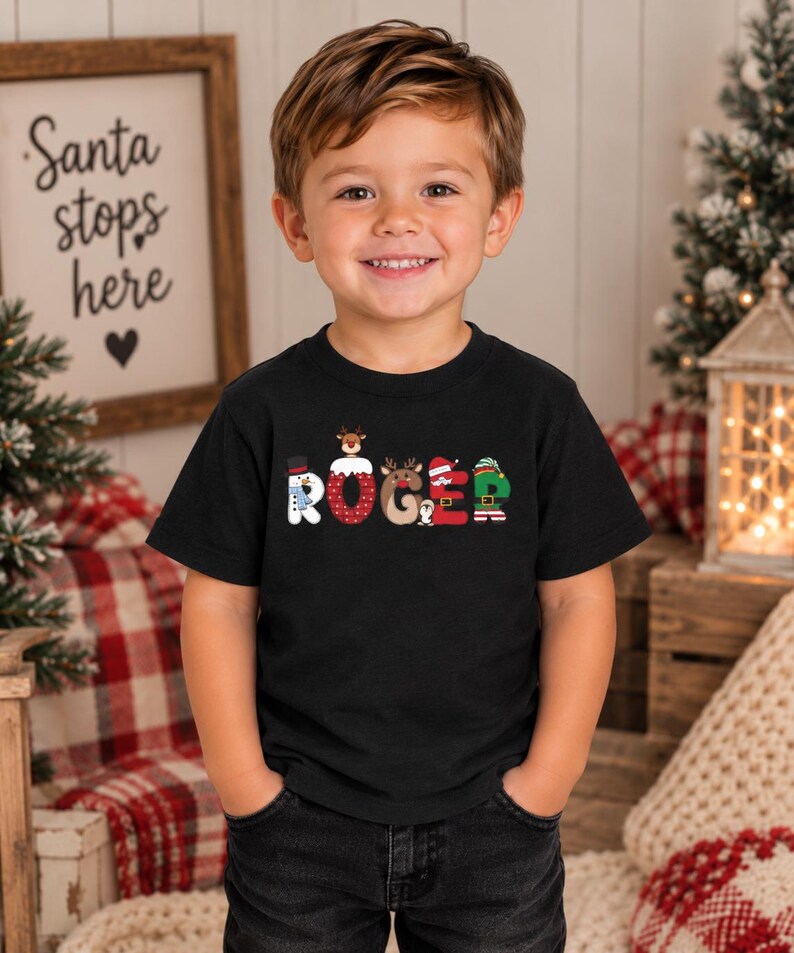 May include: A black short-sleeved t-shirt featuring the name ROGER printed in large, colorful letters. Each letter is decorated with Christmas-themed graphics, including a snowman, a reindeer, a penguin, and an elf-inspired design.