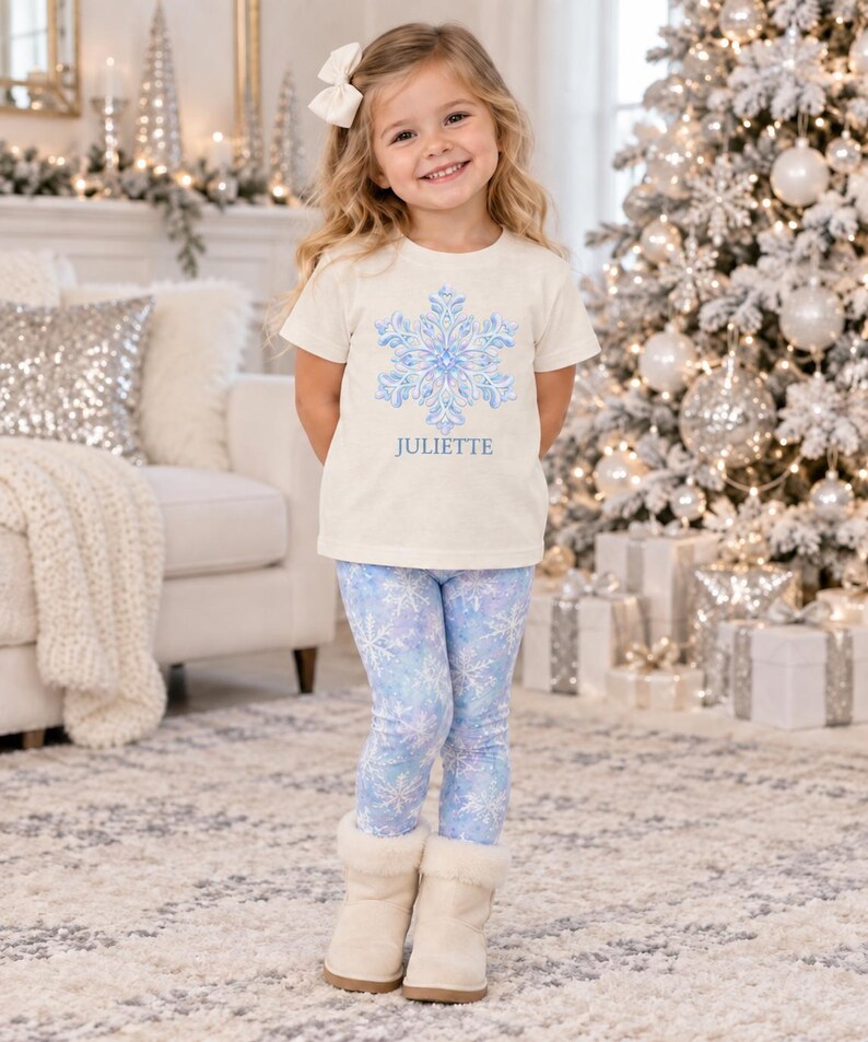 May include: A cream-colored short-sleeve t-shirt featuring a blue and white snowflake graphic with the name JULIETTE printed below it, paired with light blue leggings decorated with a white snowflake pattern and beige faux-fur lined boots.