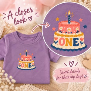 May include: A purple t-shirt featuring a graphic of a two-tiered birthday cake with the word ONE printed on the bottom layer. The cake is decorated with white flowers, a single lit candle on top, and surrounded by small colorful stars.