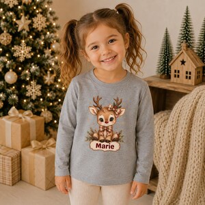 May include: A long-sleeved heather gray shirt featuring a graphic of a cartoon reindeer with a red nose, a plaid bow, and a plaid necktie. Below the reindeer, a decorative label contains the name Marie printed in a dark red script font.