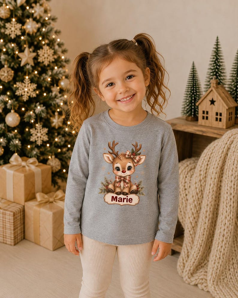 May include: A long-sleeved heather gray shirt featuring a graphic of a cartoon reindeer with a red nose, a plaid bow, and a plaid necktie. Below the reindeer, a decorative label contains the name Marie printed in a dark red script font.