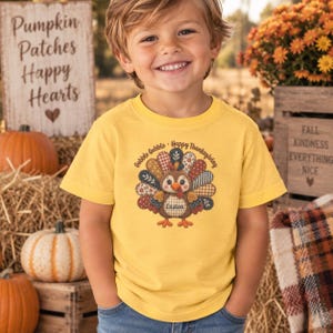 May include: A yellow short-sleeve t-shirt featuring a graphic of a turkey with a patchwork-style tail in shades of brown, blue, and cream. The text "Gobble Gobble Happy Thanksgiving" arches over the turkey, and the name "Easton" is printed on its body.