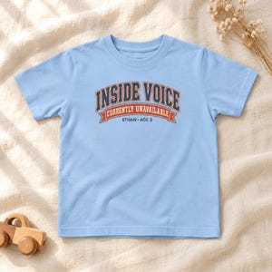 May include: A light blue short-sleeve t-shirt featuring a graphic with the text "INSIDE VOICE" in dark blue, "CURRENTLY UNAVAILABLE" in white on a red banner, and "ETHAN &bull; AGE 3" in dark blue lettering centered on the chest area.